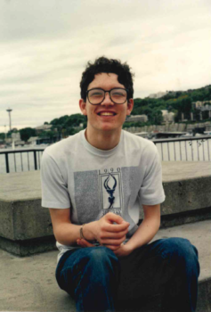 A teen boy smiles at the camera while sitting on a bench with a lake and the Space Needle in the background