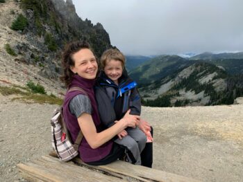 A mother and a son sitting on her lap smile at the camera while sitting on a bench