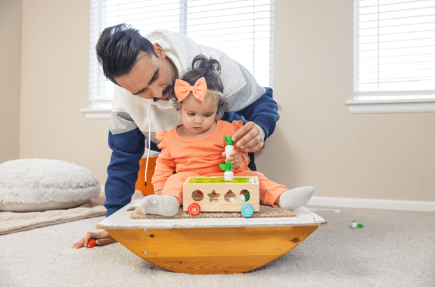 A dad plays with his little girl who is sitting on a balance board