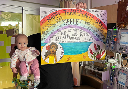 An infant is held up next to a hand-drawn Happy Transplant Day Seeley sign in her hospital room