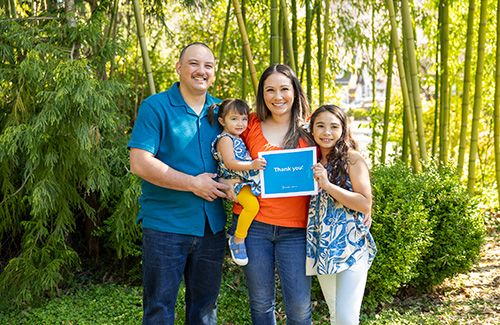 A father, mother and their two daughters hold up a teal sign with white letters that says. "Thank you!"