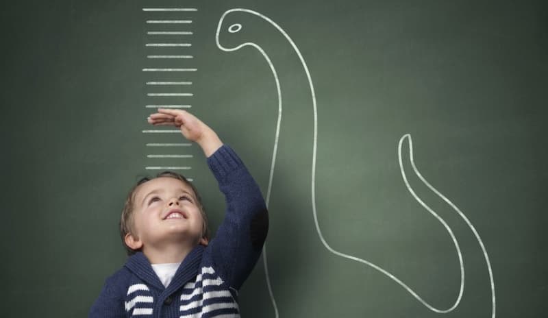 A boy measures his height next to a dinosaur drawn on a chalkboard.