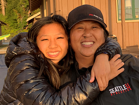 a daughter wraps her arms around her mother as they smile at the camera with the glow of a sunset lighting up their faces