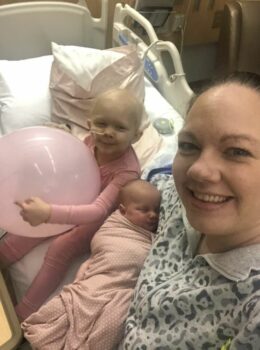 A little girl with her mom and little sister smile at the camera while sitting on her hospital bed