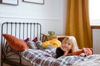 A blond school-aged girl lays on plaid comforter on her bed 