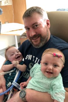 a smiling dad holds his twin infants in the hospital