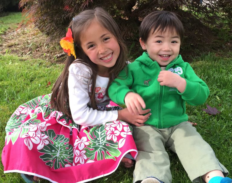 a young sister and brother smile at the camera while sitting in the grass