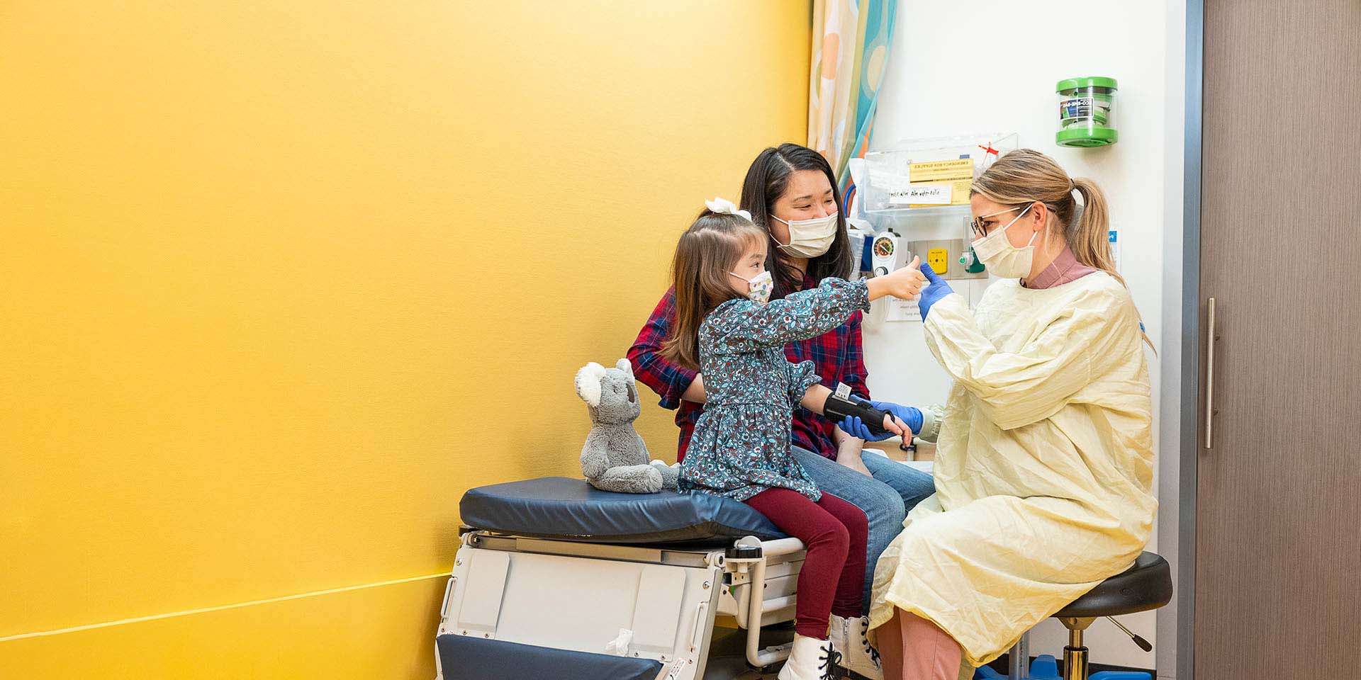 A girl gives a thumbs up to her doctor while her mother looks on