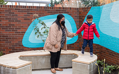 A mother holds her son's hand as he walks across a bench