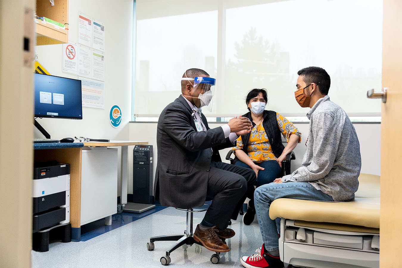 : A doctor talks with a teenage patient and their caregiver in an exam room.