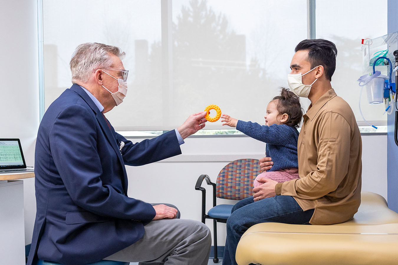 A doctor hands a toy ring to a toddler in her father's lap