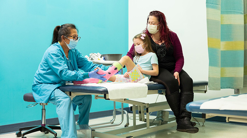 a female doctor examines foot and ankle casts on a young female patient who is laying on the exam table with her mother supporting her from behind